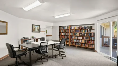 A meeting room or library with a long table, office chairs, a large bookshelf filled with books, and a sliding glass door
