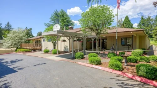 A wide view of a tan building's covered entrance with an American flag, a parking lot, and a manicured lawn